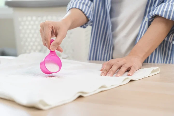 Dłoń sypiąca proszek do prania z różowej miarki na biały materiał, druga dłoń go wygładza