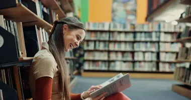 Uśmiechnięta młoda kobieta siedzi w bibliotece i czyta książkę.